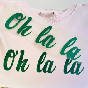 Może przedstawiać: Jasnoróżowa bluza z napisem "Oh la la" w żywym zielonym skrypcie z przodu. Bluza jest złożona, ukazując okrągły dekolt i miękki materiał.