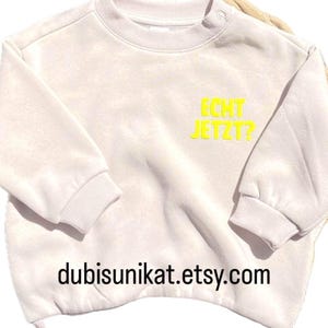 Peut inclure: Un sweat-shirt beige clair avec l'inscription "ECHT JETZT?" en jaune. L'adresse Etsy "dubisunikat.etsy.com" est imprimée en bas. Le sweat-shirt a un col rond et des manches longues.