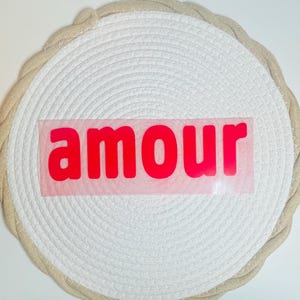 Op de afbeelding: Een ronde, wit geweven onderzetter met een beige rand. Het woord "amour" is in felrood over het midden van de onderzetter gedrukt. De onderzetter is gemaakt van een dik, gestructureerd materiaal.