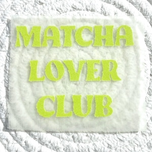 Könnte beinhalten: Ein quadratischer, hellfarbener Aufnäher mit den Worten "Matcha Lover Club" in leuchtend grünen, stilisierten Buchstaben. Der Text ist auf dem Aufnäher zentriert, der ein leicht strukturiertes Aussehen hat. Der Hintergrund ist ein weißes, gewebtes Material.