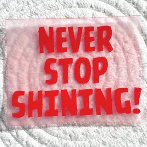 Op de afbeelding: Een rechthoekige, transparante sticker met de woorden "NEVER STOP SHINING!" in vette, rode letters. De sticker is geplaatst tegen een witte, getextureerde achtergrond.