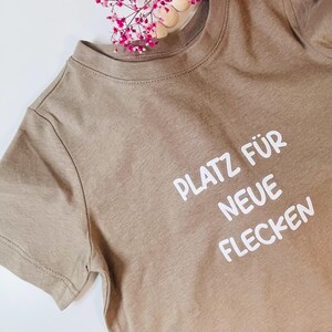 Könnte beinhalten: Ein hellbraunes T-Shirt mit dem deutschen Text "PLATZ FÜR NEUE FLECKEN" in Weiß gedruckt. Das Shirt hat kurze Ärmel und einen Rundhalsausschnitt. Rosa Blumen sind im Hintergrund.