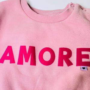 Könnte beinhalten: Hellrosa Sweatshirt mit dem Wort "AMORE" in leuchtend pinkfarbenen Blockbuchstaben auf der Vorderseite. Das Sweatshirt hat einen Rundhalsausschnitt und Druckknöpfe an der Schulter. Ein kleines Etikett mit einem Herzen ist unten rechts am Wort "ORE" befestigt.