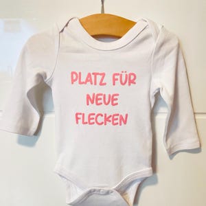 Op de afbeelding: Witte babybody met lange mouwen, hangend aan een houten hanger. De body heeft de roze tekst "PLATZ FÜR NEUE FLECKEN" op de voorkant. Het kledingstuk is ontworpen voor baby's en peuters.