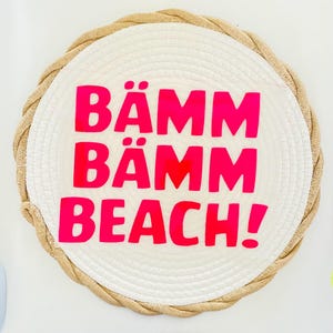 May include: A round, white woven placemat with a thick, tan rope border. The words "BÄMM BÄMM BEACH!" are printed in bright pink across the center of the placemat.