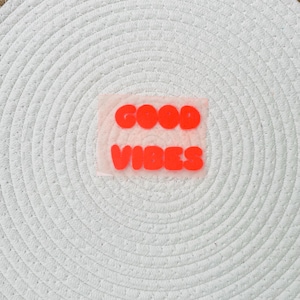 Peut inclure: Gros plan sur un dessous de plat blanc tissé avec un motif en vinyle thermocollant rouge et orange qui se lit "GOOD VIBES". Le texte est en gras, police sans-serif.