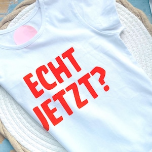 Könnte beinhalten: Weißes T-Shirt mit dem Aufdruck "ECHT JETZT?" in leuchtendem Rot. Das Shirt besteht aus weichem Material und hat einen Rundhalsausschnitt. Der Text ist in einer fetten, serifenlosen Schriftart gehalten.