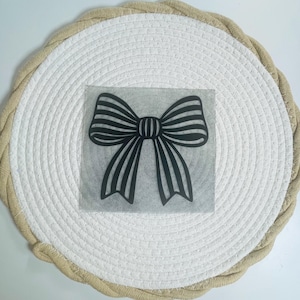 Pode incluir: Um autocolante de vinil preto com um design de laço estilizado com riscas verticais. O laço está centrado num quadrado de fita de transferência transparente, apresentando uma silhueta gráfica limpa com laços simétricos e duas pontas.