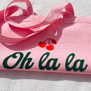 Könnte beinhalten: Eine rosa Tragetasche mit passenden Griffen. Die Tasche zeigt ein Paar rote Kirschen und die Worte "Oh la la" in grüner Schrift. Die Tasche ist aus einem weichen Stoff gefertigt und eignet sich zum Tragen von Gegenständen.
