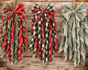 May include: Three decorative fabric bows with frayed edges hanging vertically. The first two bows feature red and green plaid patterns, while the third bow is made of sage green fabric with white stripes and small cross-shaped patterns.