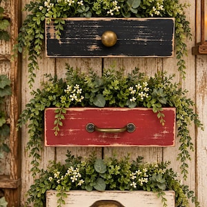 May include: Three stacked rectangular wooden drawers mounted vertically, each featuring a distressed finish in black, red, or cream. Each drawer contains artificial green foliage with small white berries and includes a metal handle or knob.
