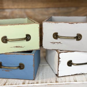 May include: Four small, square wooden boxes in pastel colors: light green, blue, gray, and white. Each box has a distressed finish and a bronze-colored handle. The boxes are stacked and arranged to show their fronts.