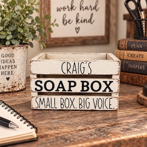 May include: A rectangular white wooden crate with a distressed finish featuring black text that reads CRAIG'S SOAP BOX on the top slat and SMALL BOX. BIG VOICE. on the bottom slat. The crate has three horizontal slats and an open top design.