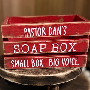 May include: A rectangular red wooden crate with a distressed finish. The front features white text that reads PASTOR DAN'S, SOAP BOX, and SMALL BOX. BIG VOICE. The crate is constructed from three horizontal wooden slats with open gaps between them.