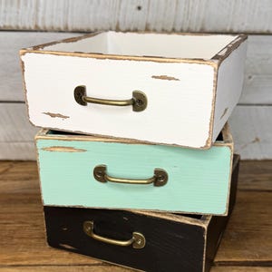 Puede incluir: Tres cajas de almacenamiento cuadradas de madera apiladas. La caja superior es blanca, la del medio es verde menta y la inferior es negra. Cada caja tiene un asa de color bronce. Las cajas tienen un aspecto rústico y envejecido.