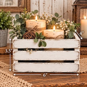 Puede incluir: Una caja de madera blanca desgastada que contiene dos velas de pilar encendidas envueltas en cordel, rodeadas de follaje verde y pequeñas flores blancas. La caja mide 15,24 cm de ancho, 8,89 cm de alto y 10,16 cm de profundidad.
