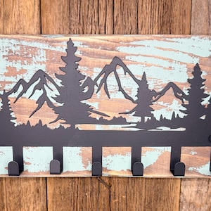 May include: A rustic key holder featuring a black metal mountain and tree silhouette design. The wooden base has a distressed teal and brown finish, with six black hooks for keys or small items.