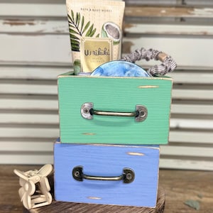 May include: A stacked set of two small wooden drawers, one green and one blue, each featuring a dark metal handle. A Bath & Body Works Waikiki Beach Coconut tube, a blue circular object, and a patterned scrunchie sit inside, with a beige hair clip nearby.