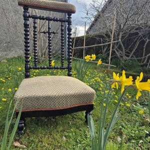 Peut inclure: Une chaise de prière en bois noir avec un siège et un dossier rembourrés. La chaise a un motif de croix au dos et est décorée de perles. La chaise est placée dans un espace herbeux avec des fleurs jaunes.