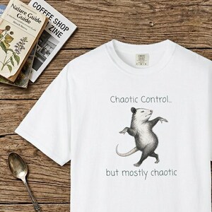 Könnte beinhalten: Ein weißes T-Shirt mit kurzen Ärmeln und einer zentralen Grafik eines stehenden Opossums. Über dem Opossum steht der Text "Chaotic Control..." und unter dem Opossum steht der Text "but mostly chaotic" in einer einfachen, dunklen Schrift.
