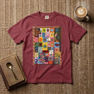 Puede incluir: Una camiseta de manga corta color granate con un gráfico rectangular central compuesto por una colorida cuadrícula de patchwork con diversas ilustraciones pequeñas, incluyendo una mariquita, un gato, motivos florales y formas geométricas.