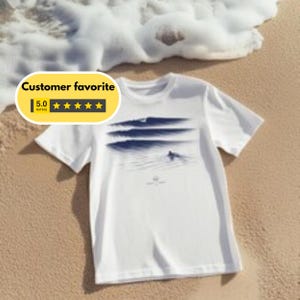 Może przedstawiać: Biały t-shirt z krótkim rękawem z ciemnoniebieskim nadrukiem graficznym przedstawiającym fale oceanu i surfera. Wzór jest wyśrodkowany na klatce piersiowej, ukazując spokojną nadmorską scenerię z warstwowymi wzorami fal i małą postacią na desce.