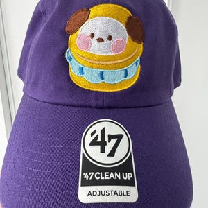 May include: Purple adjustable baseball cap with a cartoon character patch. The patch features a yellow, white, brown, and pink design. The cap has a curved brim and a tag that reads "47 Clean Up Adjustable".