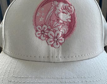Puede incluir: Gorra de béisbol beige claro con un diseño rosa bordado de un perfil femenino dentro de un marco circular, rodeado de estrellas y flores. La gorra tiene una visera curva y un diseño cosido.