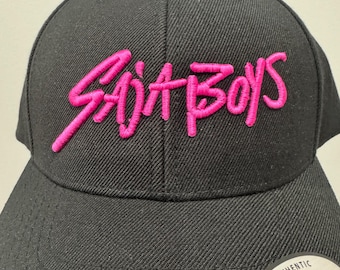 Puede incluir: Gorra snapback negra con el texto bordado "SAJIT BOYS" en rosa brillante. La gorra tiene una corona estructurada y una visera curva. Una etiqueta circular con el texto "THE AUTHENTIC YUPOONG SNAPBACK" es visible.