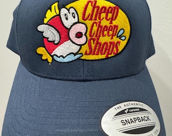 Puede incluir: Gorra snapback azul marino con un diseño bordado. El diseño incluye un pez de dibujos animados con detalles en rojo, blanco y amarillo, y el texto "Cheep Cheep Shops" en amarillo. La gorra tiene una visera plana y cierre a presión.