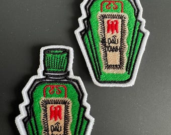 Puede incluir: Dos parches bordados con forma de botellas verdes con borde blanco. Cada parche presenta un emblema rojo y texto en un rectángulo beige. Las tapas de las botellas son verdes. Los parches están sobre una superficie gris oscuro.