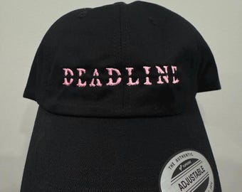 Puede incluir: Gorra de béisbol negra con la palabra "DEADLINE" bordada en rosa en la parte delantera. La gorra tiene una visera curva y una correa ajustable. Una pegatina plateada y blanca en la visera dice "ADJUSTABLE" y "YUPOONG SINCE 1974."