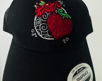 Puede incluir: Gorra snapback negra con un diseño bordado en rojo, blanco y verde. El diseño presenta una fruta roja en forma de corazón con un patrón de remolino, texto rojo y un fondo circular blanco. La gorra tiene una visera curva y cierre snapback.