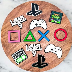 May include: A wooden circle decorated with gaming-themed shapes and text. Features PlayStation symbols, controller designs, and the phrases "Happy B-Day", "Level 9", and "Gaming Mode On". The shapes are in different colours.
