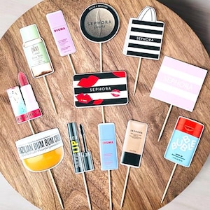 May include: A selection of cosmetic product cutouts on wooden sticks, including lipstick, eyeshadow, and skincare items. The cutouts feature the Sephora logo, along with the brands Pixi, Byoma, and Sol de Janeiro. The items are arranged on a wooden surface.