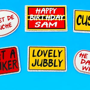 May include: A selection of stickers with humorous phrases and text, set against a blue backdrop. The stickers feature phrases such as "Bonnet de Douche," "Lovely Jubbly," and "Cushti."