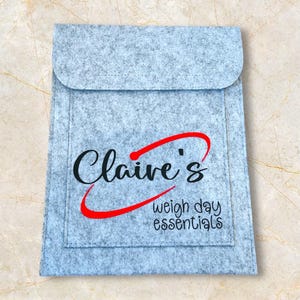 May include: A pale grey felt pouch with a curved flap. The text "Claire's weigh day essentials" is displayed in black and red script on the front.