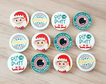 May include: A collection of twelve round Christmas-themed buttons. The buttons feature elf faces, the text "Elf on Duty", and the phrase "Santa's Approved Nice List". The buttons have red, green, and white colors.