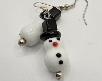 May include: A pair of snowman earrings. Each earring features a white snowman with a black top hat, black button eyes, and a red carrot nose. The earrings have silver-coloured hooks and accents.