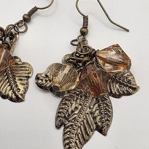 Puede incluir: Un par de pendientes de hojas de color bronce. Cada pendiente presenta un dije de hoja detallado, adornado con cuentas facetadas de color ámbar y cuentas redondas más pequeñas. Los pendientes tienen un cierre de gancho.