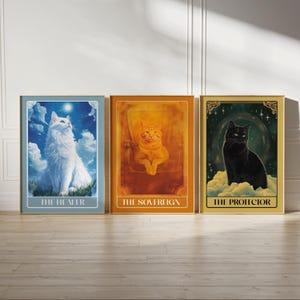 Op de afbeelding: Drie ingelijste prints met kattenillustraties en titels. De eerste print toont een witte kat met blauwe ogen die omhoog kijkt naar een halve maan. De titel is "De Genezer". De tweede print toont een oranje kat die op een troon zit. De titel is "De Soeverein". De derde print toont een zwarte kat die op een wolk zit. De titel is "De Beschermer".