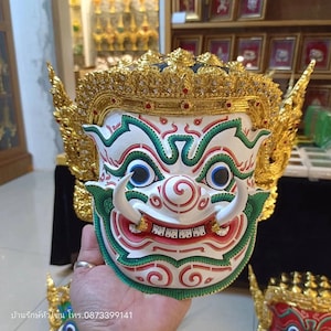 Pode incluir: Uma máscara branca, verde e vermelha com uma coroa dourada. A máscara tem uma expressão feroz com olhos grandes, uma boca larga e dentes afiados. A coroa é decorada com joias douradas e vermelhas.