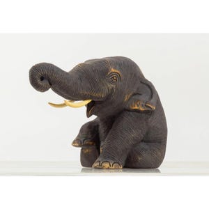 Op de afbeelding: Een donkerbruin olifantenbeeldje in zittende houding met de slurf omhoog gekruld. De olifant heeft een textuur in de huid, zichtbare ogen en twee gebogen, crèmekleurige slagtanden die uit de mondstreek naar buiten steken.