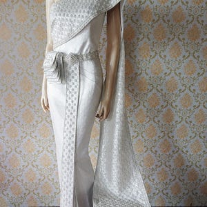 May include: A shimmering silver one-shoulder dress featuring intricate embroidery and a matching wide belt with a pleated side detail. The garment includes a long, flowing fabric panel draped over the shoulder, showcasing a textured metallic finish.