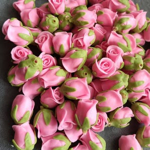 May include: A pile of small, pink rose buds with green sepals. The roses are tightly closed, suggesting they are fresh or recently crafted. The image is a close-up, highlighting the delicate details of the petals and leaves.