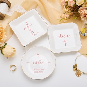 Pode incluir: Três pratos de anel de cerâmica branca com acabamento dourado. Cada prato tem uma cruz rosa e um nome, "Olivia", "Sophia" e "Camille", com uma data. Os pratos estão sobre uma superfície branca.