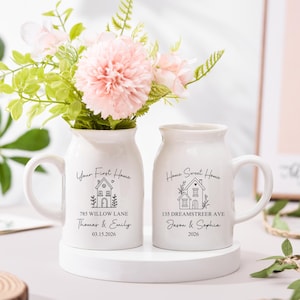 May include: Two white ceramic vases with handles, each featuring a house illustration and text. One reads "Your First Home" with an address and names, the other says "Home Sweet Home" with an address and names. Pink flowers and greenery are in the vases.