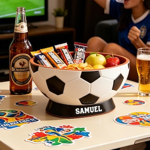 May include: A soccer ball-shaped snack bowl filled with candy bars, chips, and fruit. A bottle of beer and a glass of beer are on the table. The bowl has the name "SAMUEL" on the base. Colorful stickers are scattered on the table.