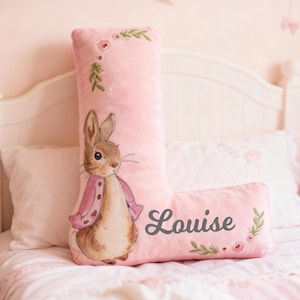 May include: A pink, plush letter "L" cushion featuring a watercolour illustration of Peter Rabbit in a pink jacket. The cushion is personalised with the name "Louise" and floral accents. It rests on a bed with white bedding.
