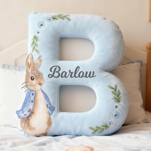 May include: A light blue, plush letter "B" pillow with the name "Barlow" in gray script. The pillow is decorated with floral and leaf accents. A watercolor illustration of Peter Rabbit is on the left side of the pillow.